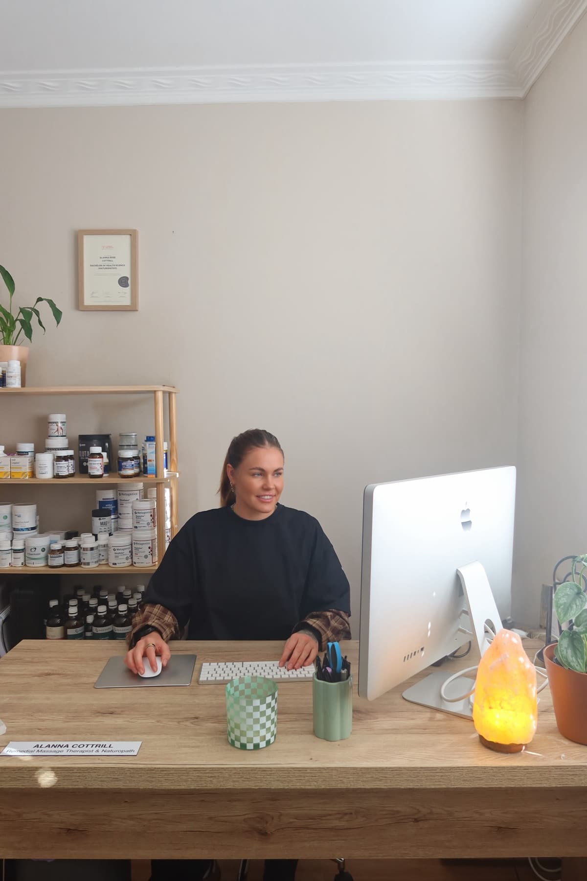 Alanna Cottrill at the clinic desk, her naturopathy degree framed on the wall behind her and shelves of herbal and nutritional medicine alongside.