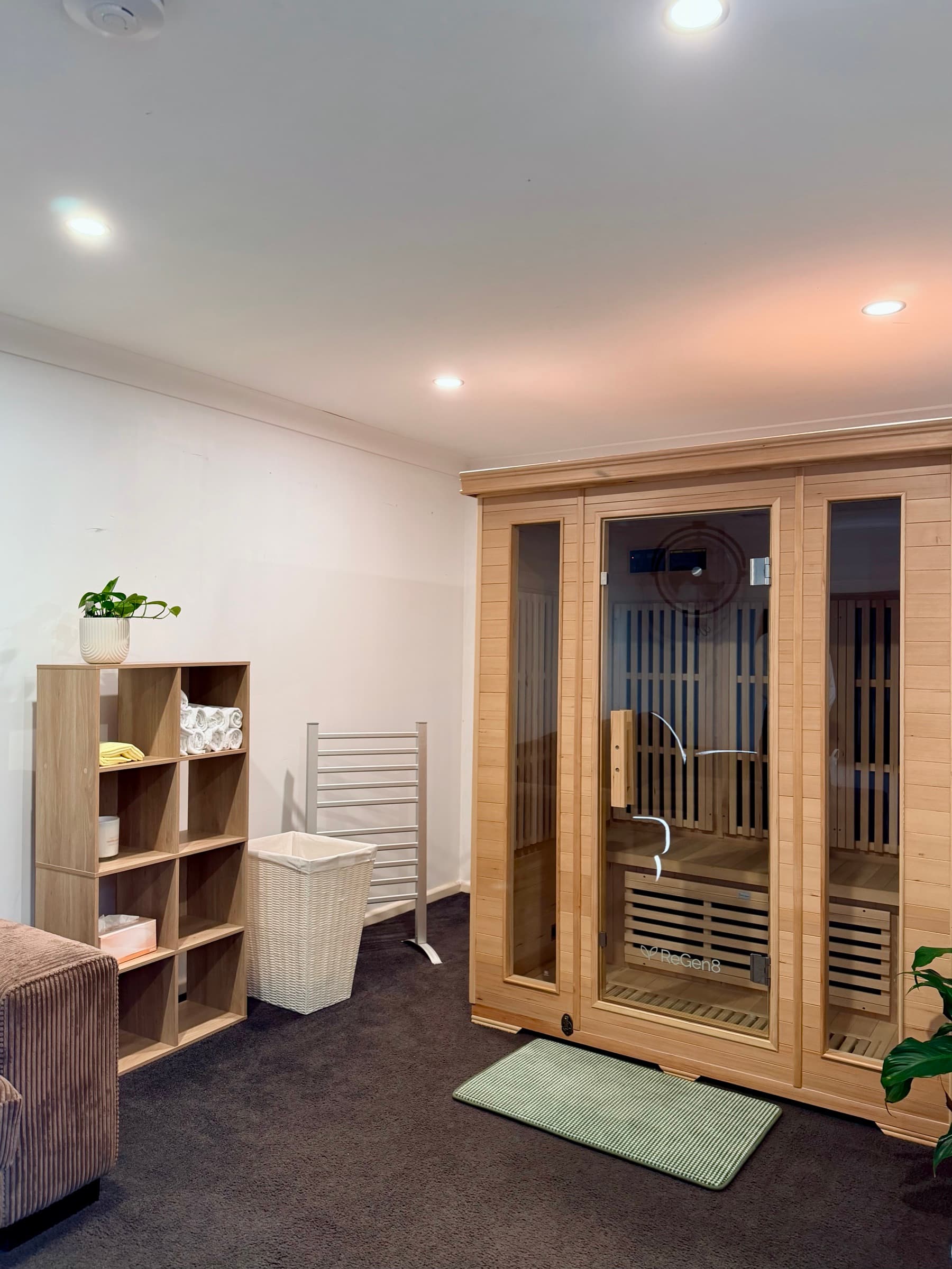 The infrared sauna at The Natural Path Collective — a timber cabin with glass doors, beside a shelf of rolled white towels and a potted plant.